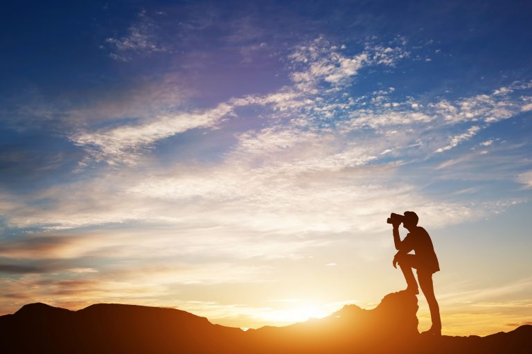 Man standing at the top of a mountain looking through looking forward through binoculars